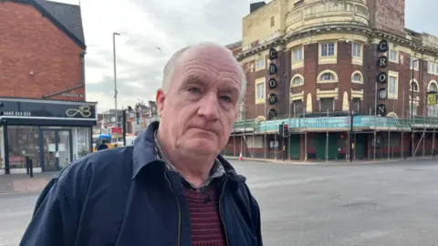 Ray Casey is standing outside the former Crown cinema in Middlesbrough which is a dilapidated red-brick building covered in scaffolding. He is wearing a blue jacket and stripey top and has white hair. He is not smiling 
