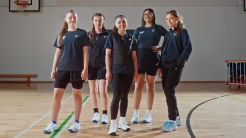ASICS A group of five girls standing in a school sports hall. There are basketball hoops and a trolley in the background. Each girl is wearing a different outfit with different parts of the kit, including black leggings, a black gilet, a navy blue half-zip jumper and black jogging pants. 
