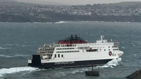 BBC The Manxman - and large white, red and black ferry with Steam-Packet.com on the side - sailing out of Douglas Bay in shoppy seas with Onchan Head in the background.