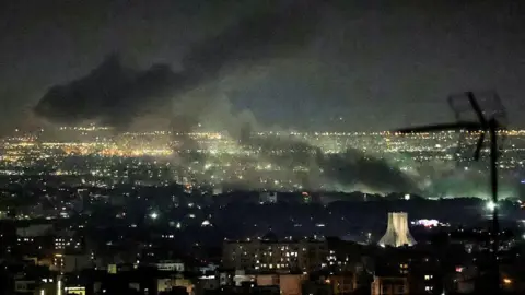Smoke plumes billow from the site of airstrikes near Azadi Tower in western Tehran on 10 March. The night sky and smoke are black. Below, lights are on in buildings and a tower is lit up.