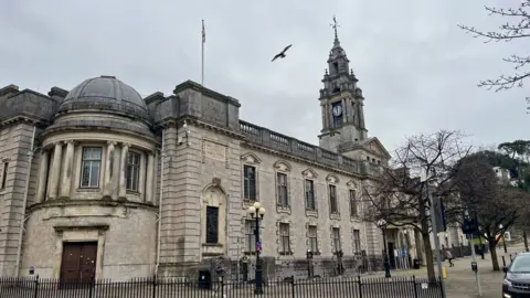 Torquay Town Hall, the headquarters of Torbay Council. The light stone building has a round part with dome on top in the foreground and then a rectangular paty with five sets of two windows which is attached to a clock tower in the background of the image. The cloudy sky is slghtly overcast and a seagul in flight is visible in the middle of the image.