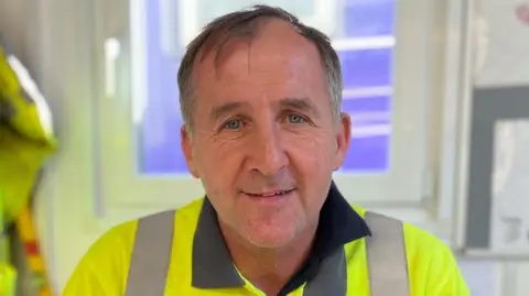 Man in a high-visibility yellow Taylor Wimpey shirt sits in an office chair, smiling at the camera, with a window, hanging work jacket, and site plans visible in the background.
