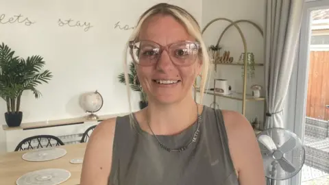 A young woman with blonde hair smiles at the camera. She is wearing pink glasses and a grey sleeveless top.