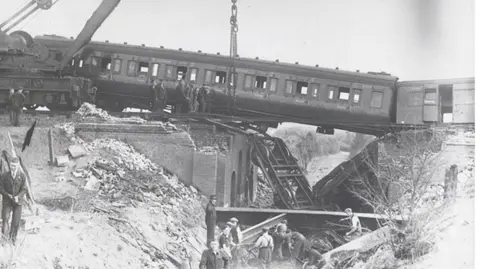 A black and white photo shows a steam train carriage suspended by a crane over a ravine. The mangled wreckage of the railway track on the bridge lies below the carriage. Men stand in the wreckage searching for survivors.