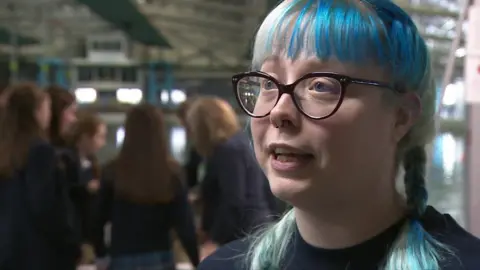 Kate Mace is looking at the camera, has dark framed glasses, a nose ring and blue hair in two plaits is wearing a dark t-shirt.