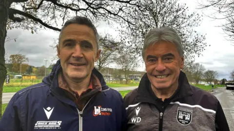 Rhys (left) and Wynne (right) both stand in a car park outside the neighbouring rugby and football clubs. Rhys has short grey hair and wears a navy raincoat with a "clwb rygby rhythun" logo. Wynne also has short silver hair, which slightly flops onto his forehead. He grins using his teeth and wears a black and grey raincoat with the football logo on the right of his zip up.
