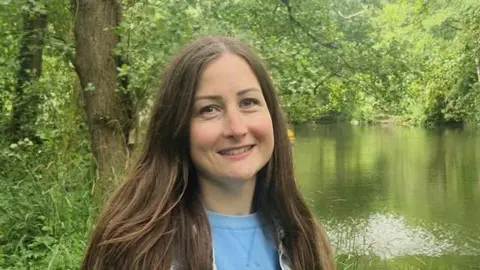 A lady with long, straight brown hair, wearing a light blue top, standing by the side of a large stream and in front of a mature tree, partly hanging over the water.