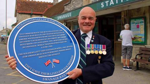 Swanage Railway Trust chairman Frank Roberts wearing a blazer bearing five medals and holding a large round blue plaque with a long inscription