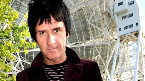 Johnny Marr, with a burgundy velvet jacket and black and white striped top, looks into the camera in front of the Lovell Telescope
