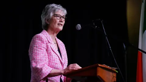 PA Media Irish president Catherine Connolly speaks during a visit to the Leeds Irish Centre, as part of her official visit to the UK. She has short white hair wearing glasses and a pink tweed jacket. She is standing at a podium giving a speech.