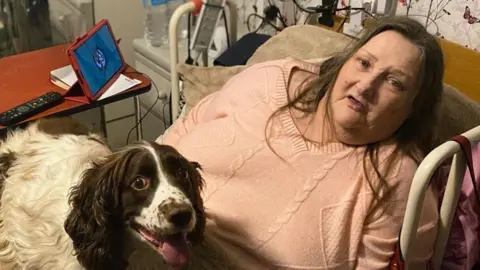 Yvonne Stride, a woman wearing a pink jumper is in bed with her dog on her lap and her i-pad with the Comfort Companions app on her bedside table.