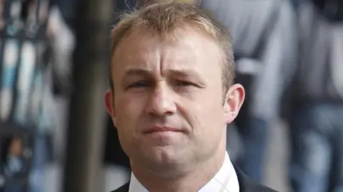 PA Media Head shot of a man with short blond hair staring blankly at the camera. He is wearing a dark suit jacket and a white shirt