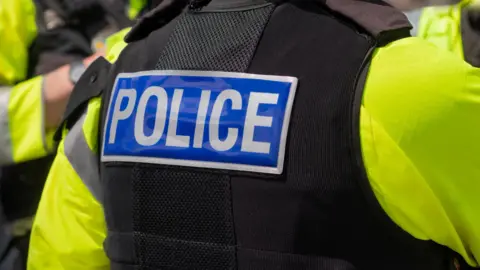 Police officer pictured from behind, wearing a high vis yellow jacket and a black vest with the word 'police' across the back. 