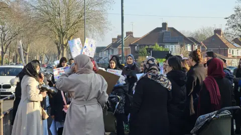 Parent Voice A group of men, women and children all wearing coats are stood at the side of the road holding placards