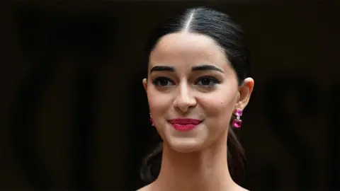 Getty Images A close up of Ananya Panday - a female actress wearing dark pink earrings, smiling. The background is black.