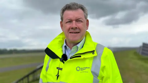 A man is wearing a yellow high-vis jacket and is standing in a field next to an airport runway. There are solar panels installed on the field.