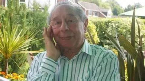 Adrian Goldberg A man sits in a garden. He has grey hair and he is wearing a green striped shirt. Trees can be seen in the background.