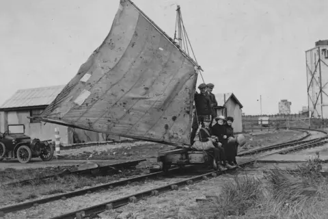Corbis via Getty Images A black and white photo showing a land yacht, a railway bogey fitted with a large sail, which runs along railway tracks. Two people are standing and two sitting on the bogey. Several buildings can be seen, along with a 1920s car.
