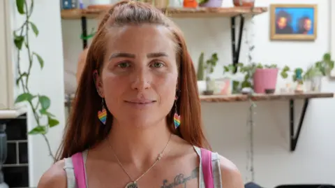 BBC Fiona is sat in the kitchen of her flat. She has longer ginger hair and the top part is pulled back. She is wearing rainbow love heart earrings and pink dungarees. In the background you can see green house plants. 