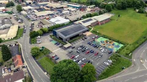 North Yorkshire Council An aerial view of the Jack Laugher Centre in Ripon, with solar panels on its roof. It has a slanted roof section in front of a flat one, and a car park around it on two sides. There's a children's play area, and to the right there are several buildings and a large expanse of green grass and trees. To the left of the building there's a large road, and behind it are industrial buildings and busy car parking.