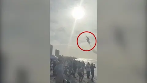 Jet flying above a crowd on a beach with a red circle around it.