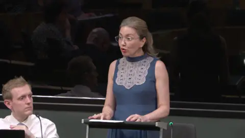 Senedd Cymru Cristiana Emsley is standing at a lectern in the Senedd. She is wearing a sleeveless blue dress with a prominent light-colored decorative lace pattern across the upper chest area. She is wearing glasses and has hair pulled back.
