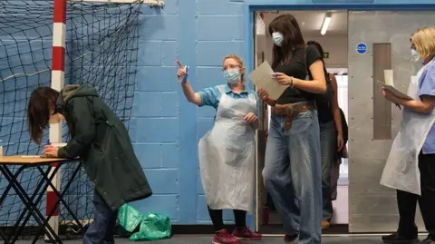 PA Media Students are walking into a sports hall carrying paperwork. Staff are near the door wearing white aprons, dealing with forms and telling people where to go. A girl in jeans and trainers and a coat is completing a form on a table near a sports hall net. The walls are blue.