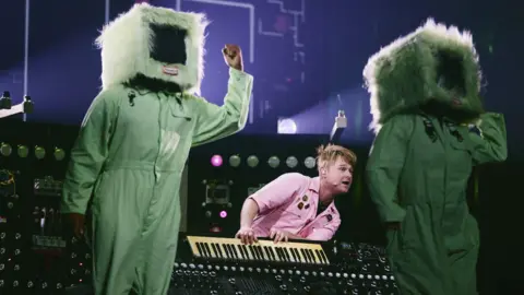 PA Media A man in a pink shirt playing a keyboard. On either side of him are two people, dressed in a green jumpsuit and a television prop over their heads.