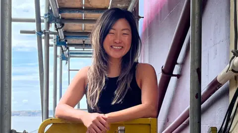 BBC A woman standing on scaffolding smiling. She is stood next to the side of a seafront pub she is painting. Behind her in the distance is the Bristol Channel.