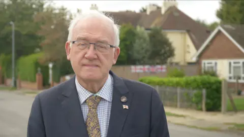 Jamie Niblock/BBC A head and shoulders image of Chris Hudson. He is wearing glasses and a blazer over a shirt and tie. He is looking into the camera.