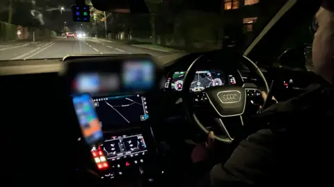 Phil Bodmer/BBC Inside of a police car seen from the back seat. The driver, on the right, gazes out of the windscreen as he travels down a dark street. In front of him, a number of screens with information are highlighted.