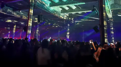 A nightclub with a crowd looking ahead to the DJ booth. Strobe lighting beams across the crowds