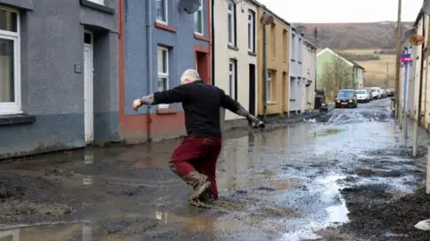 Reuters A man with grey hair, a dark top and red trousers walks to his home through deep mud covering a street in the aftermath of Storm Bert