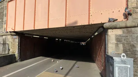 An entrance to an underpass with lots of pigeons wandering about it. There are small white dots on the ground which are droplets of pigeon poo. 