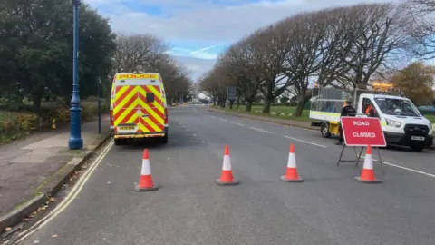 A section of road along Clarence Esplanade, there is a section of pavement to the left of the frame, which is lined with grass, trees and blue lamposts. A marked police van is parked up to the left of the road, on the right of the road is an open back truck, which a police officer wearing black uniform standing next to the driver's door. The vehicles are behind a police cordon, made using orange and white traffic cones and there is a red sign just infront of the police officer which reads 'Road closed'.