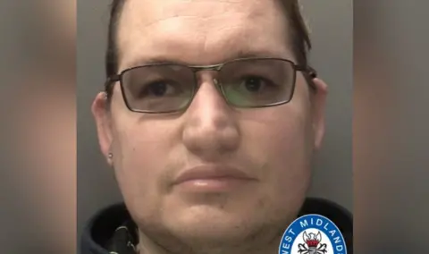 A police mugshot of a man wearing tinted black-frame glasses. He has receding dark brown hair and a stud earring in his right ear.