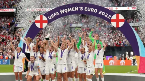 PA Media The victorious Lionesses celebrating winning the UEFA Euro 2022 on the pitch, standing in a group wearing their medals, with their hands in the air, and the captain holding the trophy aloft