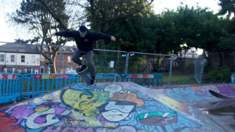 John Bray A skateboarder is wearing a black jumper, a black hat and grey jeans. He is flipping in the air on his skateboard and below him is a ramp filled with graffiti