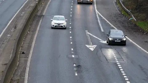 An aerial view of a motorway carriageway with three vehicles driving towards the camera. There are some potholes visible, some of which have been repaired.