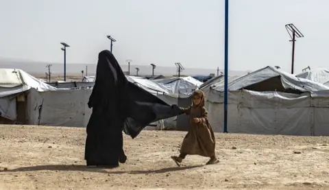 Getty Images A small child in a brown dress tugs on the back of a woman's niqab as they walk past white tents on sandy ground