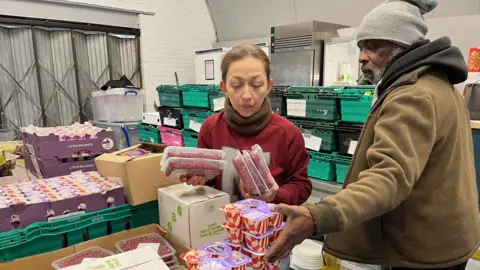 A woman in a red top and brown scarf holds packets of minced meat as a man to her rights in a grey hat, dark hoodie and light brown coat stacks pots of yoghurt on a table. Piled up green crates of food can be seen in the background