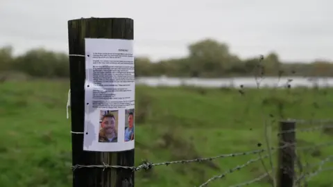 A grass area with a lake in the background.  There is a barbed wire fence with wooden posts in the foreground. One is holding an A4 notice with two pictures of Jamie Attwood.