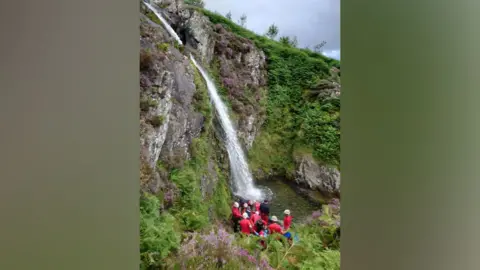 KMRT Group of rescuers in red at the bottom of a mountain waterfall, rescuing a man who has fallen from the top