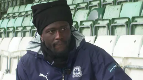 Brendan Wiredu wearing a navy coat with the Plymouth Argyle badge on it. He is sitting on a chair in the stand of a stadium.