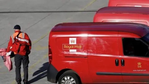 PA Media A Royal Mail worker carrying a large red mail bag walks past a number of red mail vans
