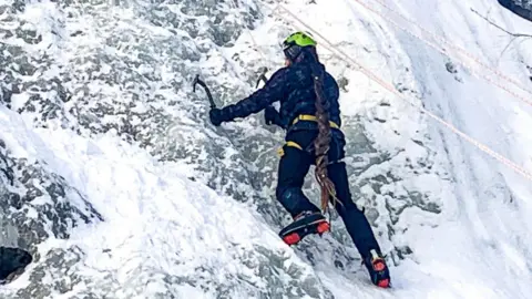 Steve Gay Steve is climbing the ice with two pick axes and his boots and is dug into the wall. He is wearing dark blue trousers and a jacket and a light green helmet. Steve has long hair in dreads which comes all the way down his back and almost to his feet.