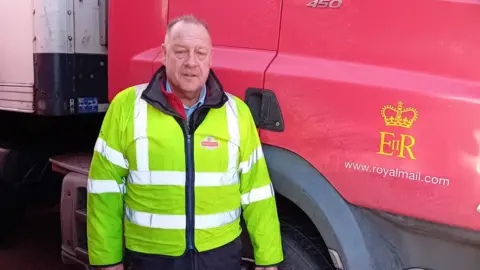 Dave Surtees Royal Mail worker Dave Surtees stands next to a Royal Mail lorry. He is wearing a high-vis jacket with the Royal Mail logo.