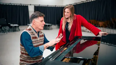 Tommy Ga-Ken Wan Ricky Ross wearing a multi-coloured sweater-vest over a blue denim shirt sitting a black piano with his hands raised level with his chest. He is seated. Next to him stands Frances Poet, wearing a red jacket over a black top. She has long, light hair which reaches her chest.