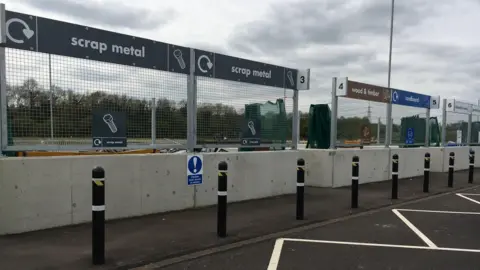 Central Bedfordshire Council Thorn Turn recycling centre, showing parking pays, several bollards, and different areas of dispose of rubbish, with trees in the far distance. 