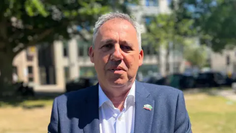 Constable Andy Jehan is a man with grey hair. He is looking into the camera. He is standing in Parade Park, with the government building out-of-focus behind him. He is wearing a grey suit and white shirt.